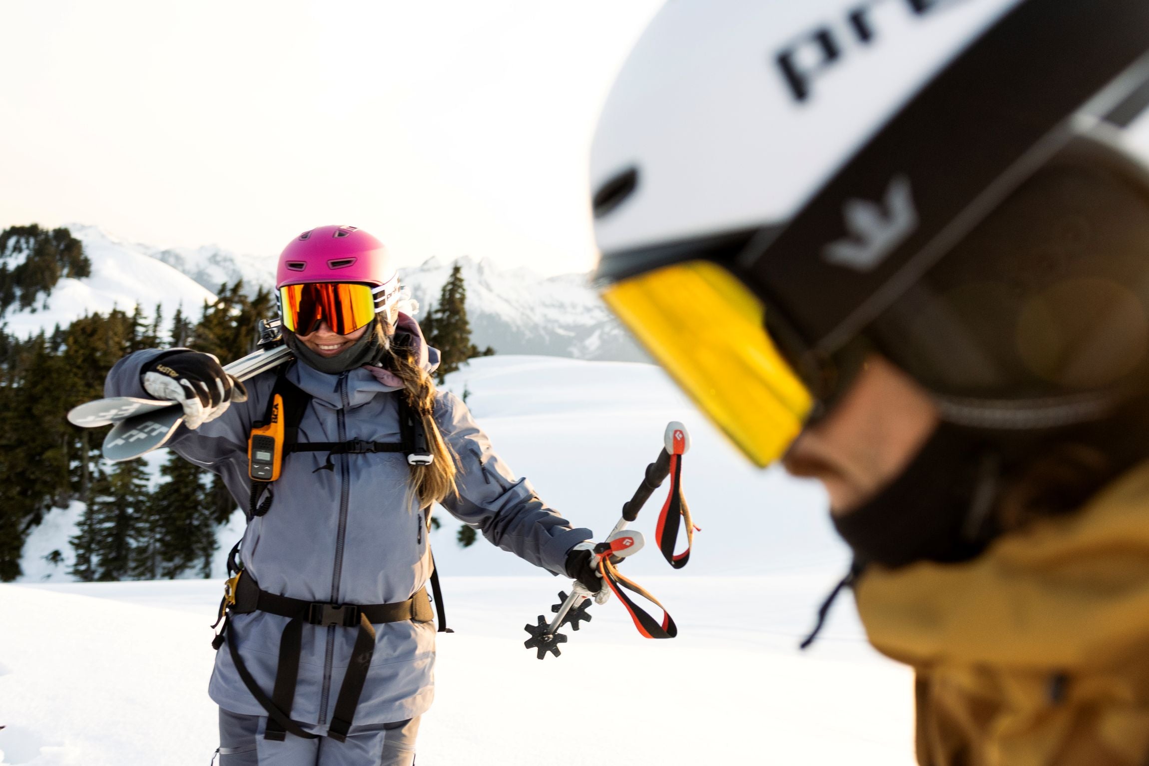 person in snowy landscape holding skis and poles and wearing helmet and goggles with another person looking down in the foreground