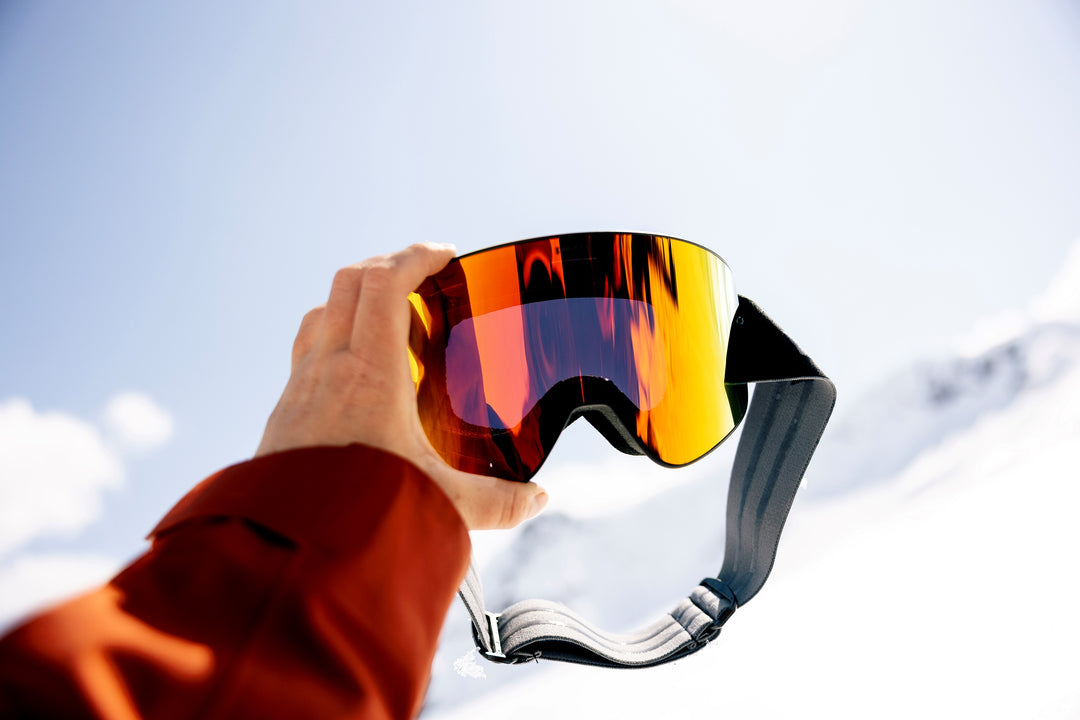 Person holding a pair of ski goggles with a snowy mountain background