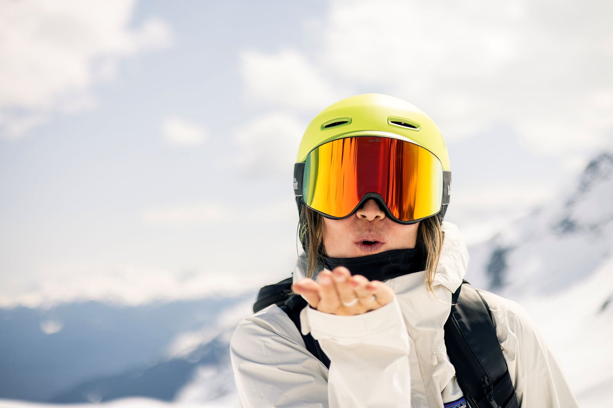woman wearing bright green ski helmet and orange goggles blowing a kiss