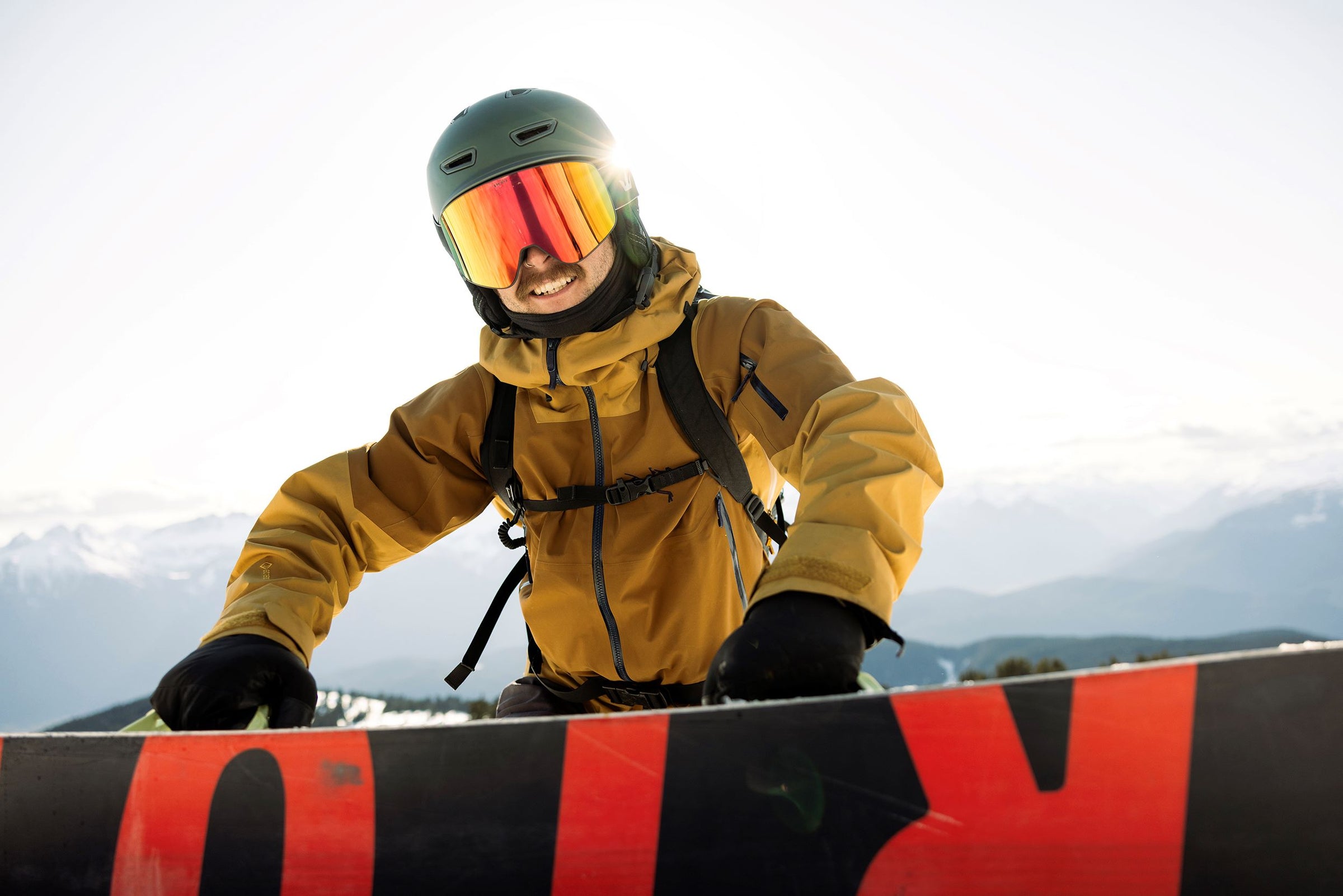 Person in yellow jacket and helmet with goggles holding a snowboard against a mountainous background