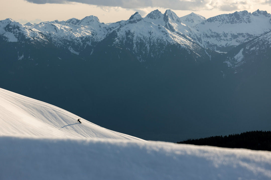 Person skiing in the distance on a snowy slope with moody snow-capped mountains in the background