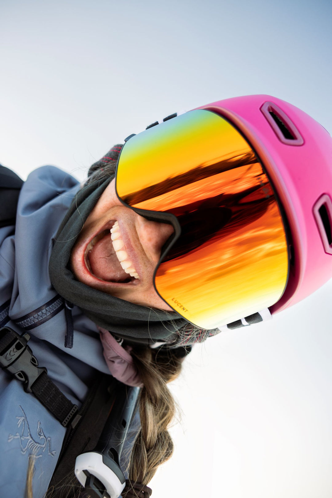 smiling person wearing a pink helmet with reflective goggles against a clear blue sky.