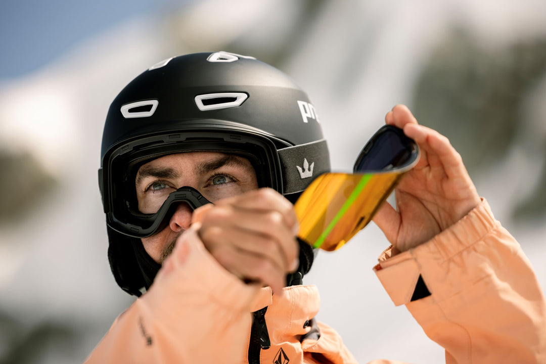 Person wearing a black helmet and orange jacket holding snapmag goggle lenses with a blurred background