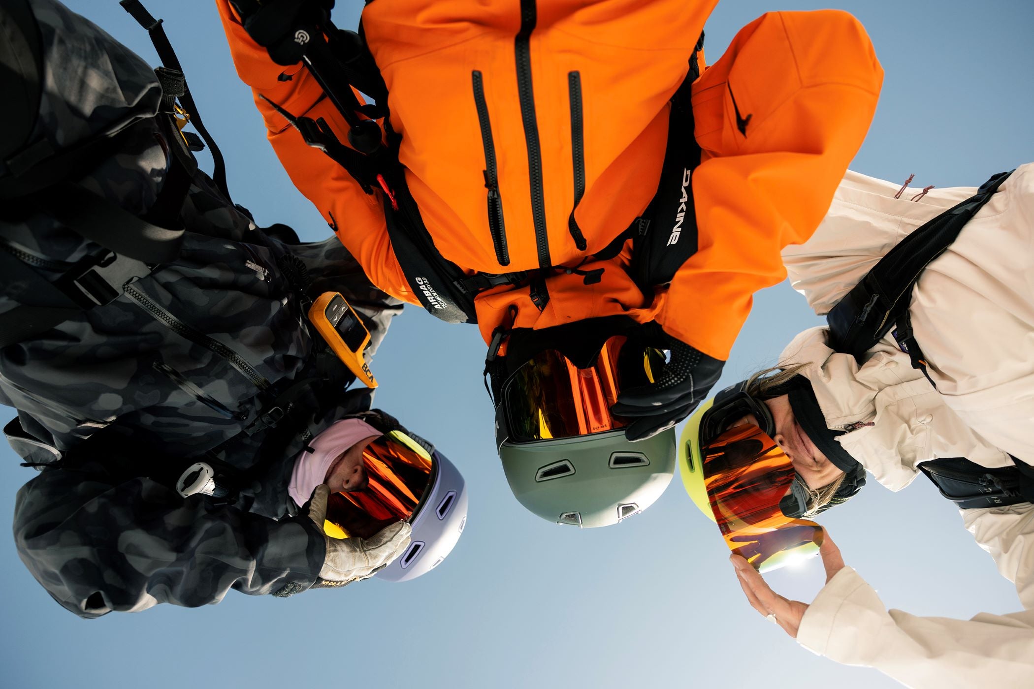 Three people in snow gear and wearing ski goggles looking down to the camera with a clear blue sky background