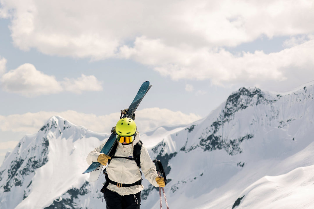 Person holding skis wearing lime ski helmet on a snowy mountain peak with clouds in the sky