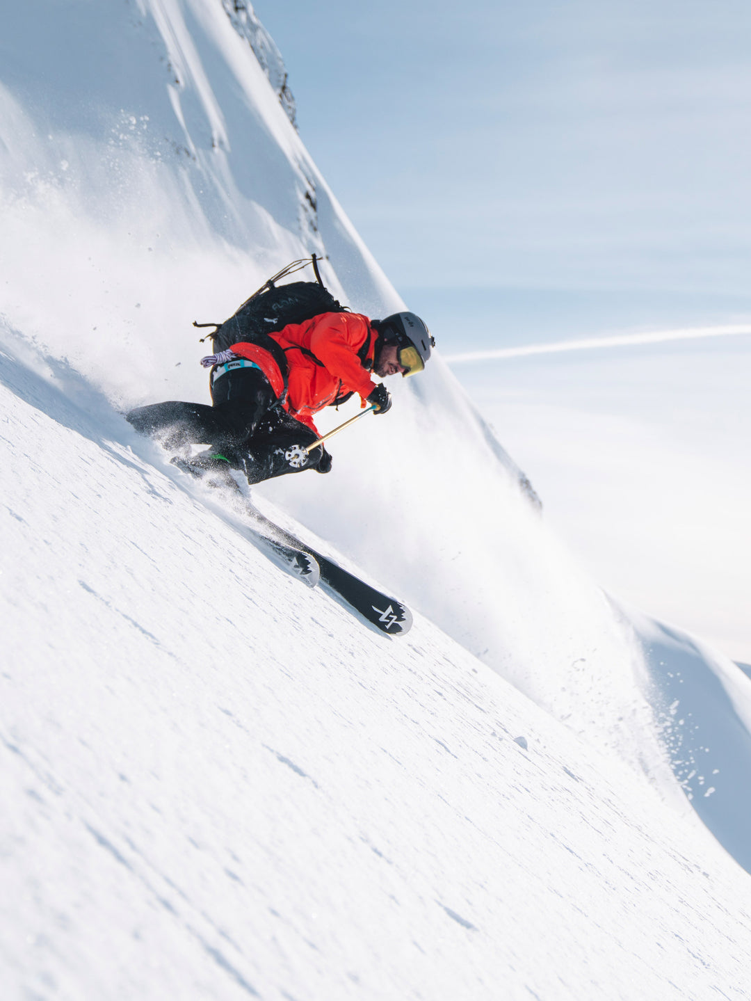 Skier in red jacket and black pants carving through powder snow on a sunny day.