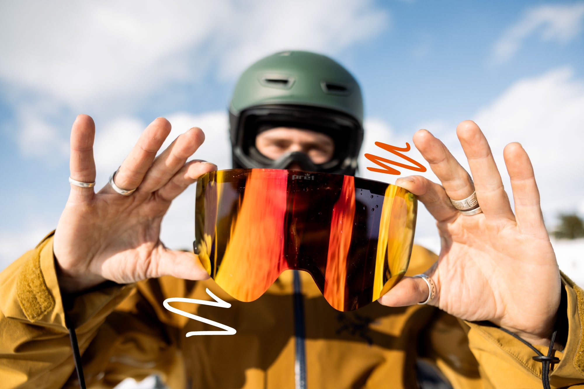 Person holding colorful goggle lense with a helmet and jacket against a blue sky.